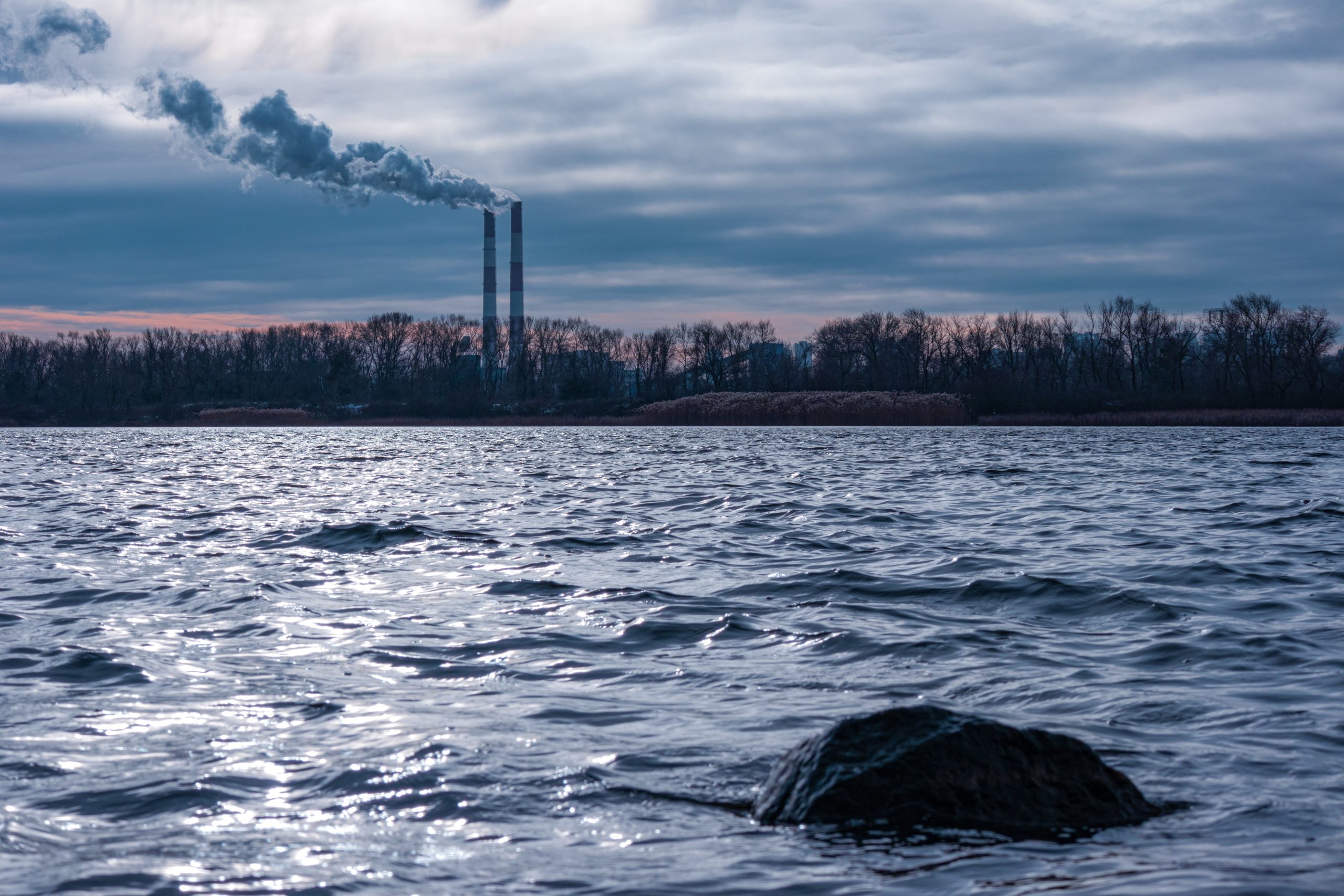 shot of a river in the foregroundwith trees and exhaust chimneys signifying emissions and investors